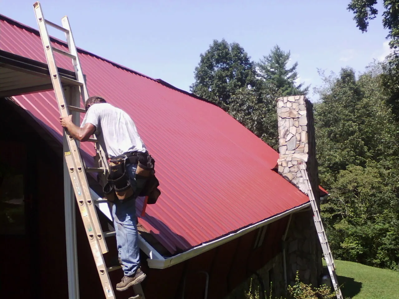Roofer on a ladder beside a red metal roof and stone chimney — Soffit & Fascia Repair by Roofing Brothers in Forestdale