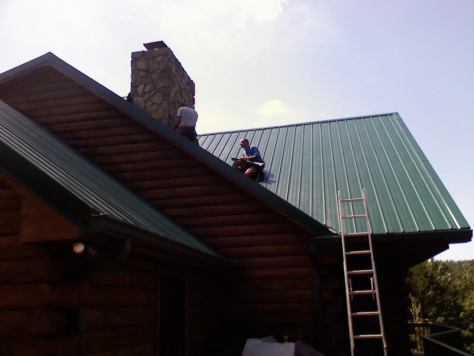 Roofers installing green metal panels beside a stone chimney on a log home in Forestdale